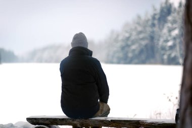 Gri bereli ve siyah ceketli bir adam, karlı bir gölün kenarındaki bankta oturuyor, kışın manzarasına bakıyor.