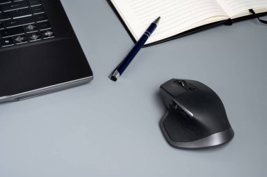 Dizüstü bilgisayar, kablosuz fare, kalem ve defter gri bir masada.