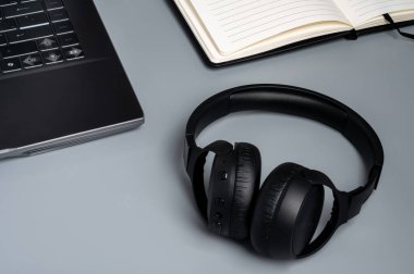 Siyah kablosuz kulaklıklar gri bir masada dizüstü bilgisayar ve açık bir defter..