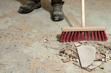 Beton zemindeki süpürge ve güvenlik botlarıyla inşaat temizliği sürüyor. Tadilat alanında iş güvenliği, el emeği, endüstriyel temizlik ve inşaat sonrası bakım kavramı