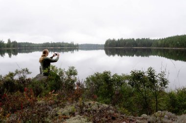 Sakin bir orman gölünün kenarında oturan kişi, sakin manzarayı akıllı bir telefonla fotoğraflıyor. Huzurlu doğa sahnesi Yansımaları ağaçlar ve yumuşak ışık
