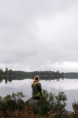 Ağaçlar ve doğal bitki örtüsüyle çevrili, bulutlu bir gökyüzünün altında sakin bir orman gölünün yanında oturan kişi. Fotokopi alanı ve sakin ruh haliyle huzurlu bir açık hava sahnesi.