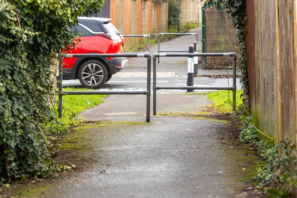 Kırmızı araba dar bir patikadan görünen ve metal bariyerlerle çevrili bir yerleşim yerinden geçiyor.
