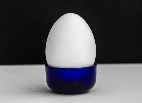 Beyaz tavuk yumurtası, koyu arkaplanı olan tarafsız bir yüzeyde, küçük, mavi bir bardağın içinde dikiliyordu. Minimal natürmort yiyecek konsepti, stüdyo ışıklandırması, fotokopi alanı ile temiz kompozisyon.