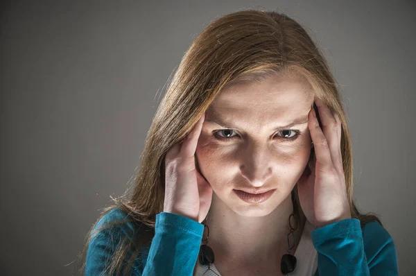 Frontal head and shoulder portrait of a young woman with a pained face looking at the camera and holding her hands on her temples against a dark gray background.