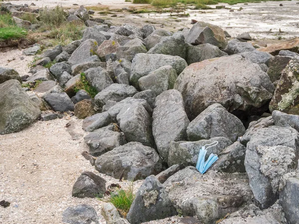 A light blue mouth and nose mask lying on boulders on the North Sea shore.
