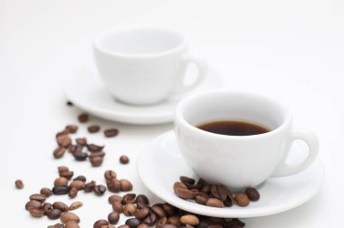 İki fincan kapuçino, çay tabağı ve beyaz arka planda kızarmış kahve çekirdekleri. Kahve içeceği dolu ön fincana odaklan..