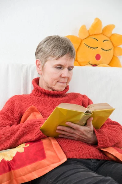 一个老年妇女在白色沙发上看书的上半身照片 在她身后的白墙上有一个日光浴状的沙发垫 — 图库照片