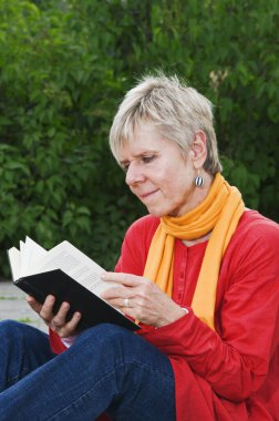 Bir parkın kenarında oturan ve önünde duran açık bir kitaba bakan olgun bir kadının yan görüntüsü.. 