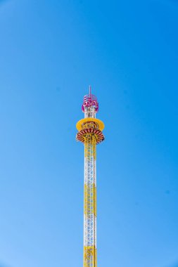 Bir lunaparkta ya da lunaparkta açık mavi gökyüzüne doğru uzun bir iniş kulesi yükselir. Parlak renkler ve minimum kompozisyon yükseklik, özgürlük ve heyecan hissi veriyor. Boş zamanları, seyahatleri ve macera temalarını resmetmek için mükemmel.
