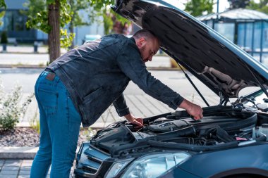 A man standing in front of a car with the hood open, inspecting the engine outdoors on a sunny day. The scene shows vehicle maintenance or troubleshooting, with the man leaning over the engine bay while checking for issues. The image illustrates