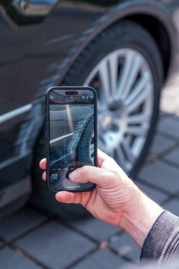 Close-up image of a man holding a smartphone and photographing a scratch on the side of a black car. The photo focuses on documenting vehicle damage for insurance, repair estimates, or accident reporting. The scratched paint and damaged area near the