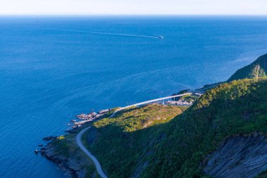 A scenic landscape featuring a winding coastal road running along steep green cliffs beside the deep blue ocean. The photo captures the serene shoreline, clear sky, and distant horizon on a sunny day, highlighting the natural beauty of the coastline.