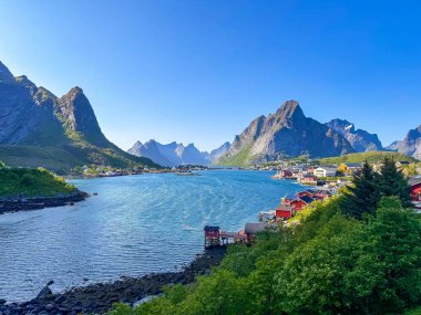 Berrak gökyüzü altında canlı kıyı limanı, demirlemiş teknelerle dolu kıyı şeridi, dramatik dağ zeminleri ve aktif demirleme, kano ve küçük yelkenli turizm enerjisi, ideal