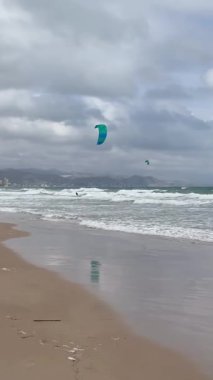 Kasvetli sahil görüntüsü. Canlı hareketli bulutlu kıyı şeridi. Enerjik rüzgar ve dalgalarla donuk plaj sahnesi. Kasvetli kıyı manzarası renkli hava akımları ve canlı su sporları sergiliyor.