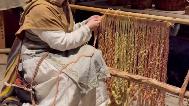 Zanaatkar kadın el dokuması dokuma tezgahında toprak rengi şal ve keten elbise giyiyor, parmaklar ahşap çerçeve üzerinde renk ipliği ve warp iplikleri ayarlıyor sabit ritmik hareketler örgülü desenler oluşturuyor