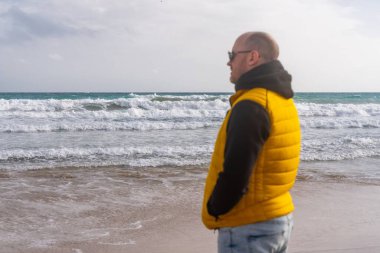 Sarı yelekli beyaz adam rüzgarlı sahil kollarında ceplerinde duruyor bulutlu gökyüzünün altındaki huzursuz denize bakıyor düşünceli gezgin yansıtıcı duraksamalar yapıyor sessiz dalgalar algılıyor.