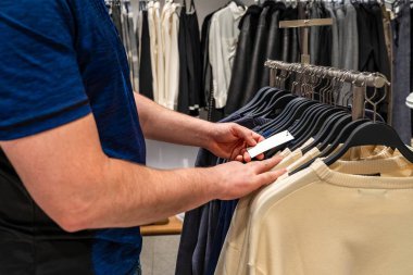 Tarayıcıda etiketi kontrol eden kişi, askıda birden fazla tepeye sahip karar anı, boyutları ve fiyatı tarayan parmaklar, sessiz renk paleti ve odaklı seçimle modern perakende iç mekan