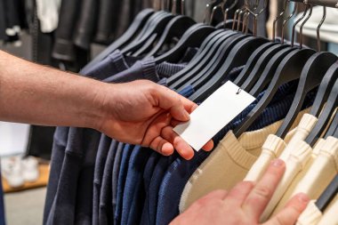 Süveterler arasındaki el karşılaştırma etiketi, kumaş ağırlığı ve rengini denetleyen stilist, pamuk örgünün dokunsal kontrolü, butik aydınlatma altında dizilmiş askılar, kalite kontrol ve seçim sahnesi