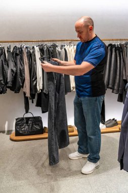 Donanma pantolonlarını butikte karşılaştıran, alışverişe odaklanmış kumaş dokusunu inceleyen, zinde ve dikişleri kontrol eden, nötr ışıklandırma altında asılı pantolon askıları, minimalist modern perakende atmosferi