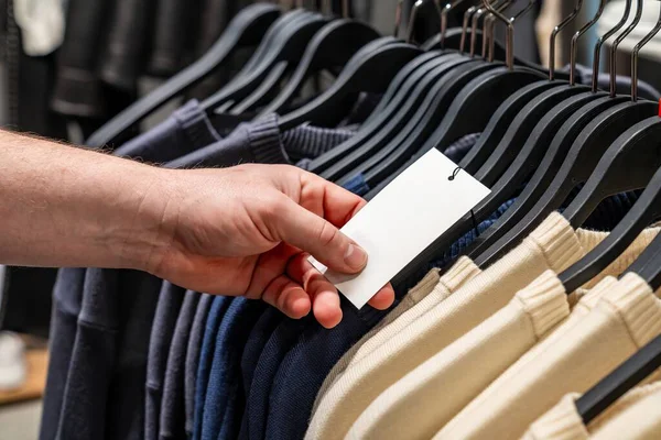 Ceketin üzerindeki fiyat etiketini inceliyor, modern butikte seçenekleri karşılaştıran bütçe bilinci olan alışverişçi, hafif ışıklandırma, askılardaki nötr kumaşlar, minimalist ekran, sessiz gezinme anı, karar verme anı,