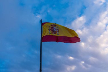 İspanyol bayrağı bulutlu gökyüzüne karşı dalgalanıyor, kıyı festivali sırasında liman direğinin üzerinde koyu renkler, rüzgârlı hareket, arka aydınlatma kenarları, vatanseverlik ve seyahat fotoğrafçılığını çağrıştıran dinamik kompozisyon