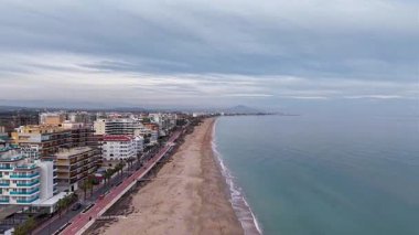 Uzun, boş deniz manzaralı cadde süpürme, İHA geniş kumlu sahil, pastel cepheler ve palmiye ağaçları arasında kıyı yolunu takip ediyor, cam su alçak bulutlarla buluşuyor, şehir için sakin bir görüntü.