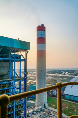 Büyük bir baca yığını gün batımında bir endüstriyel fabrikanın üzerinde yükseliyor, enerji üretimini, enerji altyapısını ve sanayi faaliyetlerinin potansiyel çevresel etkilerini vurguluyor.