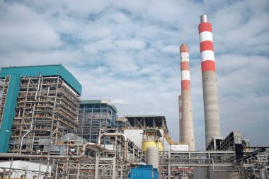 Bu görüntü, kırmızı ve beyaz çizgilerle süslenmiş uzun bacaları olan bir elektrik santralinin hakim olduğu bir sanayi sahnesini yakalıyor. Boru ve yapılardan oluşan karmaşık ağ karmaşık bir sistem olduğunu gösteriyor.