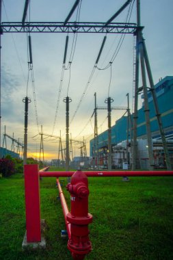 Etkileyici düşük açılı çekim, karmaşık elektrik kablolarının bitişiğindeki kırmızı yangın musluğunu vurguluyor. Arkaplan, karşıtlığı artıran ince bir gradyan gökyüzüne sahiptir.