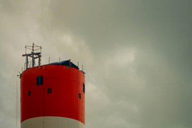 Çarpıcı kırmızı ve beyaz silindirik bir işaret ışığı dramatik bulutlu bir gökyüzünün arka planına karşı dimdik ayakta duruyor. Resim yapının yüksekliğini ve canlı renkler arasındaki kontrastı vurgular.
