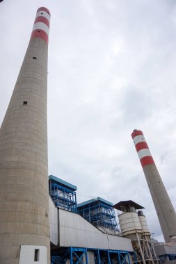 Yukarıya bakan bir perspektif, bir sanayi santralinin görkemli bacalarını yakalar. Çizgili desenleri, sessiz ve bulutlu gökyüzüne ters düşer. Beton ve çelik mimarisi...