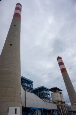 Düşük açılı bir çekimde bulutlu bir gökyüzüne karşı iki uzun baca sergilenir. Enerji üretimi ve teknolojik gelişmeyi temsil eden sanayi yapıları, araziye hakim.,
