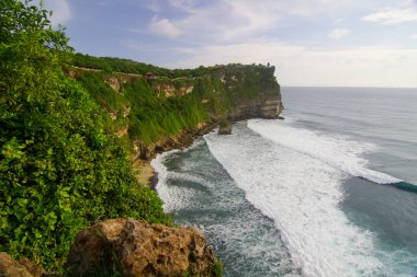 Uluwatu uçurumunun manzarası, bereketli yeşillikler, çarpan dalgalar ve engin mavi bir okyanusla dramatik kıyı şeridini gösteriyor. Tropikal manzara bir macera duygusunu davet eder.