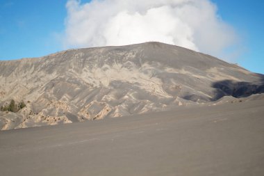 Çerçeveye hükmeden bir dağın olduğu sert bir volkanik manzara. Yukarıdaki mavi gökyüzünde, etkin volkan ortamının ham ve jeolojik gücünü vurgulayan ruhani beyaz bulutlar sürükleniyor..
