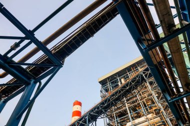 Düşük açılı bir çekim, temiz mavi gökyüzüne karşı endüstriyel bir tesiste karmaşık boru ve metal yapıları yakalıyor. Bu resim modern sanayinin karmaşıklığını ve ölçeğini gösteriyor.