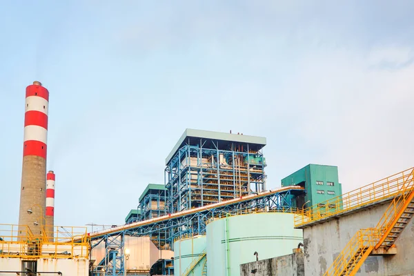 Düşük açılı çekimde, kırmızı ve beyaz çizgili, çeşitli metal yapılar ve depolama tankları bulunan sanayi kompleksi sergileniyor. Görüntü enerjinin ölçeğini yakalıyor.