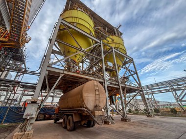 Büyük bir endüstriyel tank kamyonu karmaşık bir fabrika altyapısında depolama silolarının altına park edilmiş..