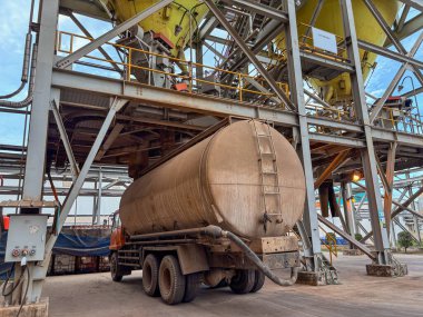 Endüstri sahnesinde, dışarıda bulunan karmaşık metal yapıya karşı kül taşıyan bir tanker görülüyor..
