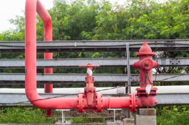 Sanayi bölgesinde belirgin bir kapakçık sistemine sahip kırmızı bir itfaiye vanası ve boru hattı altyapısı bulunuyor ve bölgeyi çevreleyen canlı yeşil bitki örtüsü bulunuyor. Sahne ruhun özünü yakalar.