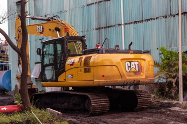 Sarı bir tırtıl kazıcısı endüstriyel bir binanın yakınındaki bir inşaat sahasının üzerine park edilmiş. Ağır makineler toprak zeminde kazı ya da inşaat için hazır bekliyor. Kazıcının.