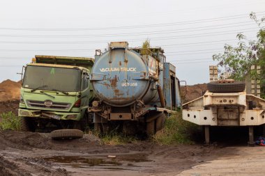 Eski ve yıpranmış ağır yük kamyonu koleksiyonunu ve çamurlu bir arazide park etmiş bir römorku gösteren bir görüntü. Kablolar gökyüzüne doğru uzanıyor. Araçlar paslanmış ve kullanılmamış görünüyor.