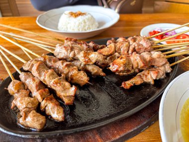 Koyu bir tabakta beyaz pirinçle sunulan lezzetli ızgara satay şişleri, yemek pişirmeyi davet eder.
