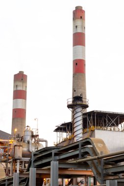 Bu resimde belirgin kırmızı ve beyaz çizgilerle süslenmiş uzun bacaların yer aldığı bir sanayi bitkisi görülüyor. Sahne endüstrinin özünü yakalıyor, çeşitli yapılar, borular ve