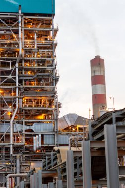 Bu görüntü, karmaşık endüstriyel altyapısını aydınlatılmış yapılar ve duman yayan önde gelen bir bacayla gösteren bir enerji santralini yakalıyor. Enerjinin kesişimini vurgular.