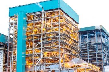 Bu görüntü, metal yapılarını ve enerji üretim sürecini aydınlatan parlak ışıkları vurgulayan bir endüstriyel kompleksin karmaşık mimarisini yakalar. Karmaşıklığı gösteriyor.