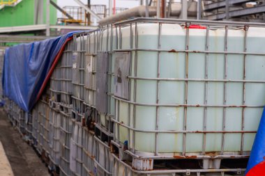 Yakın plan çekimlerde, sanayi depolama çözümleri ve lojistiğin vurgulandığı orta büyüklükteki konteynırlar (IBC 'ler) ile mavi bir muşamba sağlanarak toplu sıvıların ele alınmasında kullanılıyor.