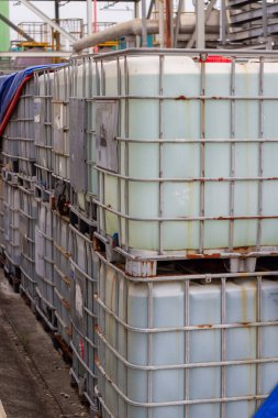 Görüntü, bir imalat fabrikasındaki endüstriyel orta büyüklükteki konteynırları (IBC 'ler) gösteriyor. Metal kafeslere kapatılmış ve paletlere yerleştirilmiş bu IBC 'ler genellikle depolama ve