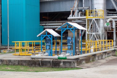 Bu görüntüde canlı mavi ve beyaz makineli bir sanayi fabrikasının küçük ve tepe çatılı çatıları altında olduğu görülüyor. Sarı güvenlik korkulukları çevreyi sararken, çeşitli borular ve yapılar örülüyor.