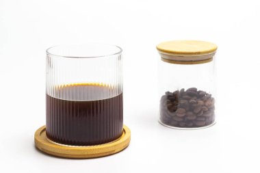 Kahvenin özünü yakalayan zarif bir hayat. Koyu kahveyle dolu bir bardak, temiz beyaz bir fonda, kavrulmuş fasulye kavanozunun yanında, ahşap bir bardak üzerinde duruyor. Kompozisyon